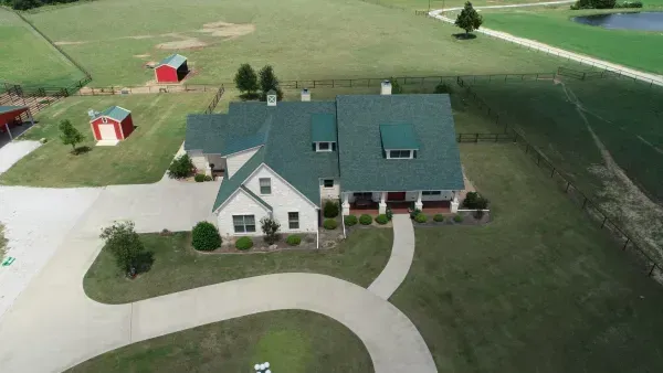Aerial view of a white house with green roof, curved driveway, lawn, and small red-outbuildings.