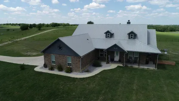 A brick farmhouse with gray roof sits on a green lawn in a rural countryside setting.