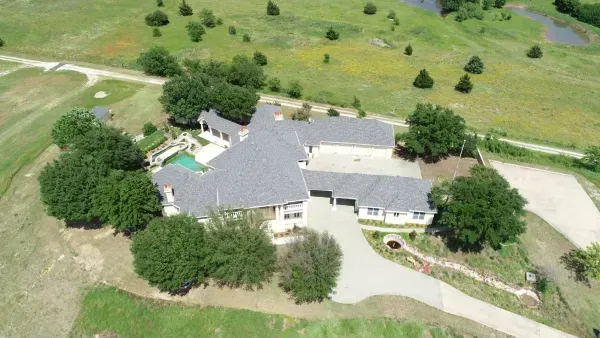 Aerial view of a large house with gray roof, driveway, and surrounding trees on a green property.