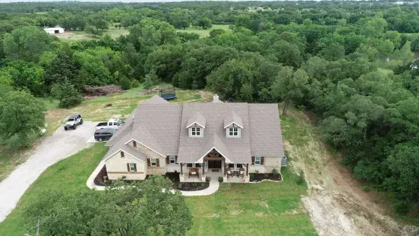 Aerial view of a large beige house with a gray roof, surrounded by dense green trees and a curved driveway.