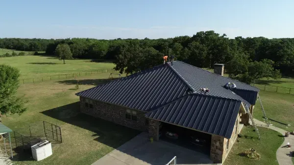 Aerial view of a stone house with a dark metal roof beside open grassy fields and trees