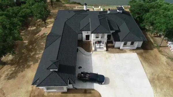 Aerial view of a large dark-roofed house with a driveway and parked car, surrounded by trees.