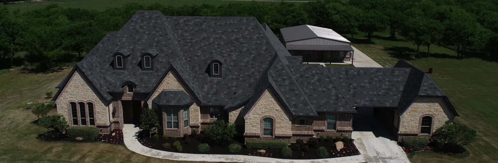 Large stone house with a dark gray roof, front lawn, and driveway in a suburban setting