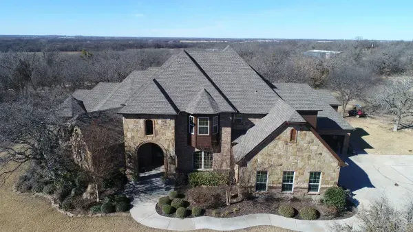 Aerial view of a large stone suburban house with a gray roof and landscaped yard
