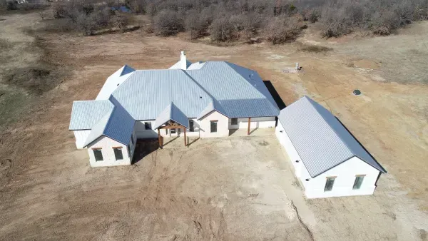 Aerial view of a large gray-roofed house and detached garage on a barren rural lot.