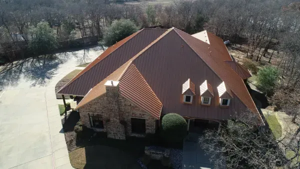 Aerial view of a large brick house with a red roof beside a pond and wooded yard