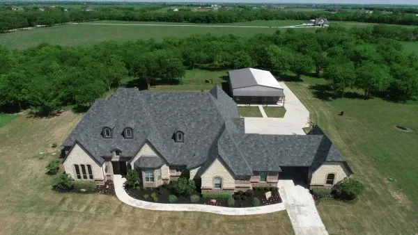 Aerial view of a large suburban house with a gray roof, front lawn, and surrounding green fields.