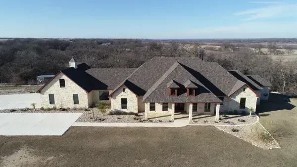 Large suburban house with stone exterior, dark roof, and a front porch on a wide lot with bare trees behind.