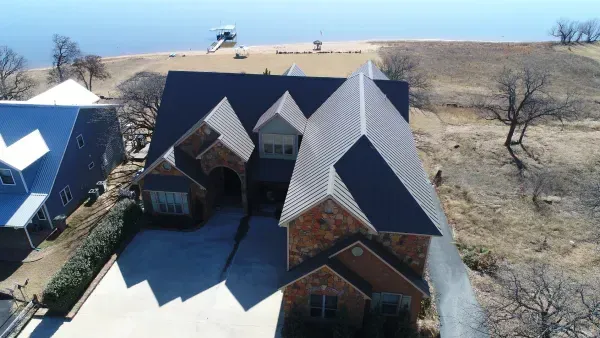 Aerial view of a large brick house with a dark roof near a grassy hillside and distant water.