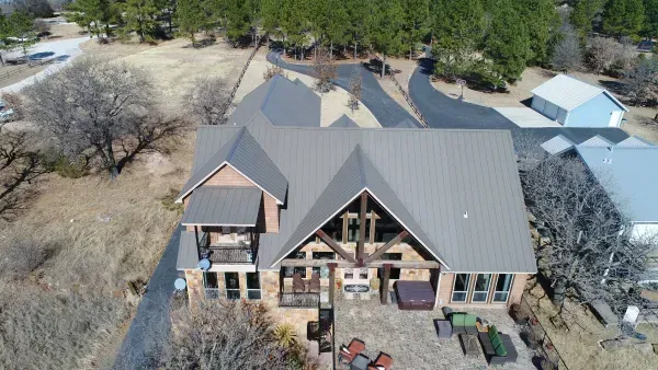 Aerial view of a rustic house with a dark metal roof and patio, surrounded by dry trees and yard.