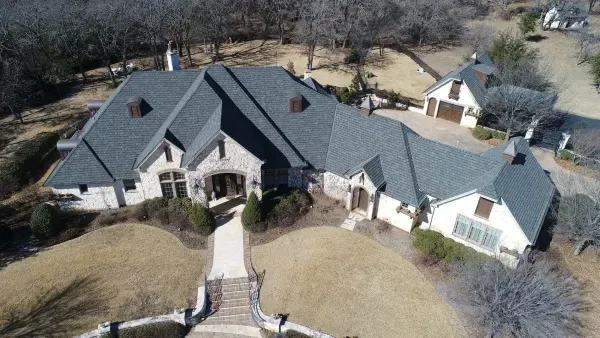 Aerial view of a large gray-roofed house with a stone entry and curved driveway on a wooded lot