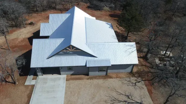 Aerial view of a white metal-roofed house with a large front driveway in a rural, wooded setting