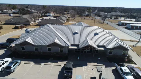 Aerial view of a large gray-roofed building with parking lot and surrounding homes on a clear day