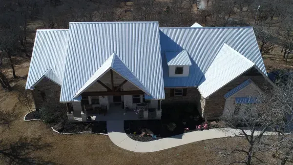 Aerial view of a house with a light metal roof, front porch, and curved driveway surrounded by trees.