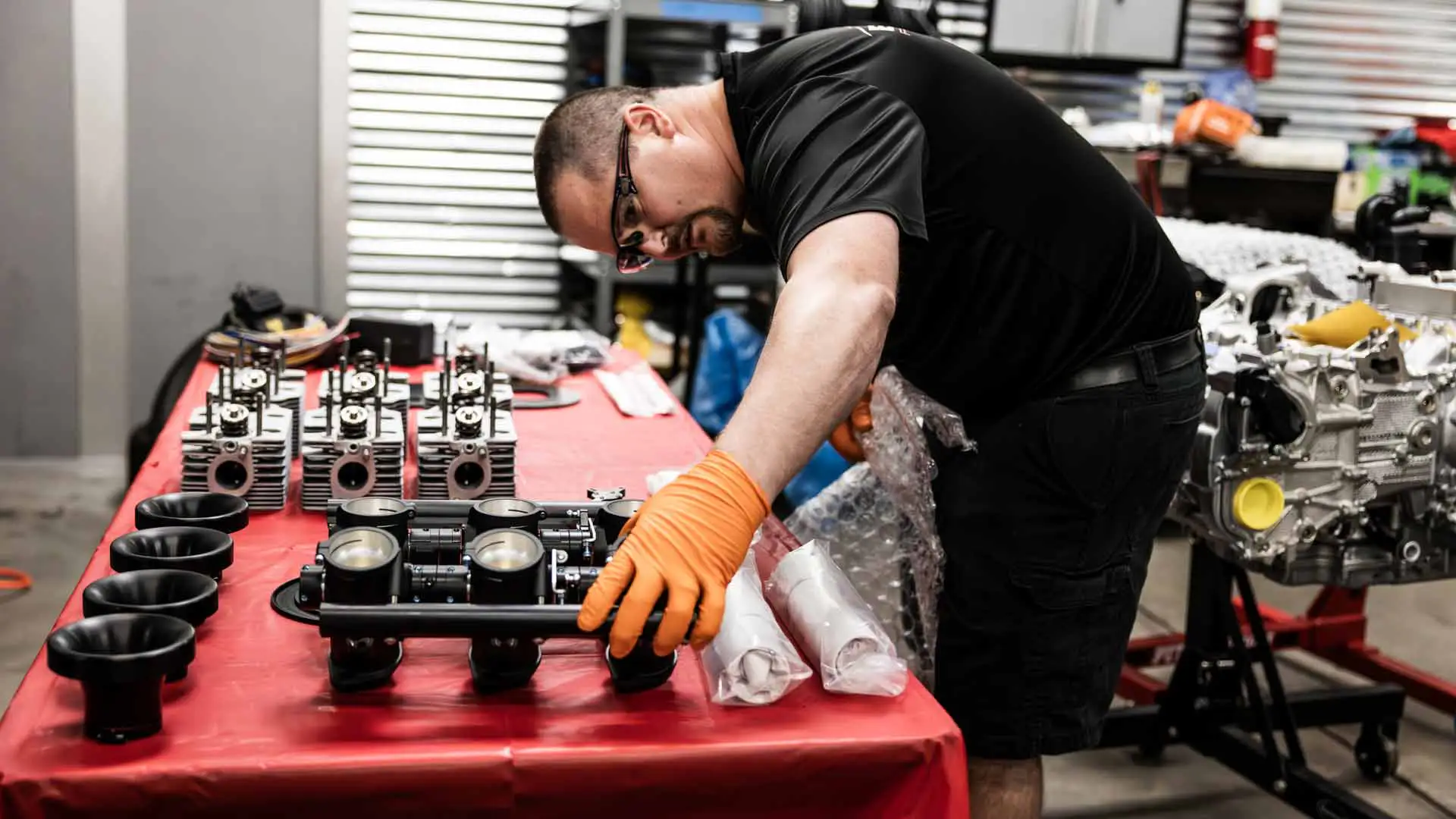 Mechanic in an auto shop wearing gloves assembles engine parts on a red table.