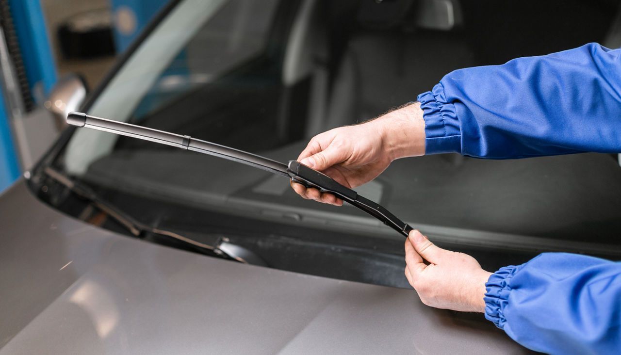 A mechanic replacing a windshield wiper blade on a gray car.