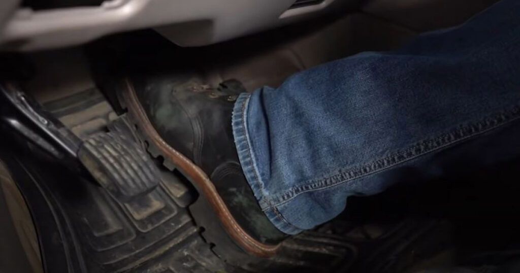 Foot pressing the brake pedal in a car, wearing a blue jeans and a black boot.