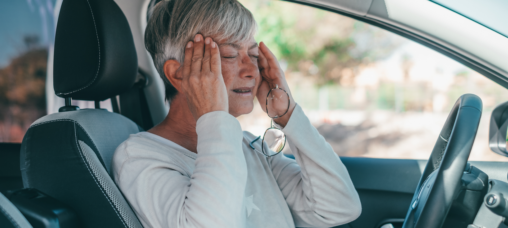 Woman in a car with hands on her head, appears to be experiencing a headache, distressed expression.