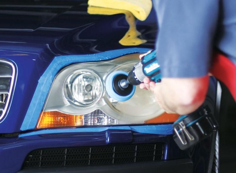 Person polishes car headlight with a power tool, blue tape outlines the work area on a blue car.