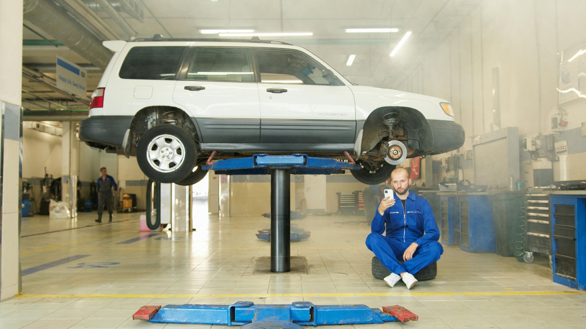 Mechanic in blue overalls sits on a tire under a lifted white car in a garage, holding a phone.