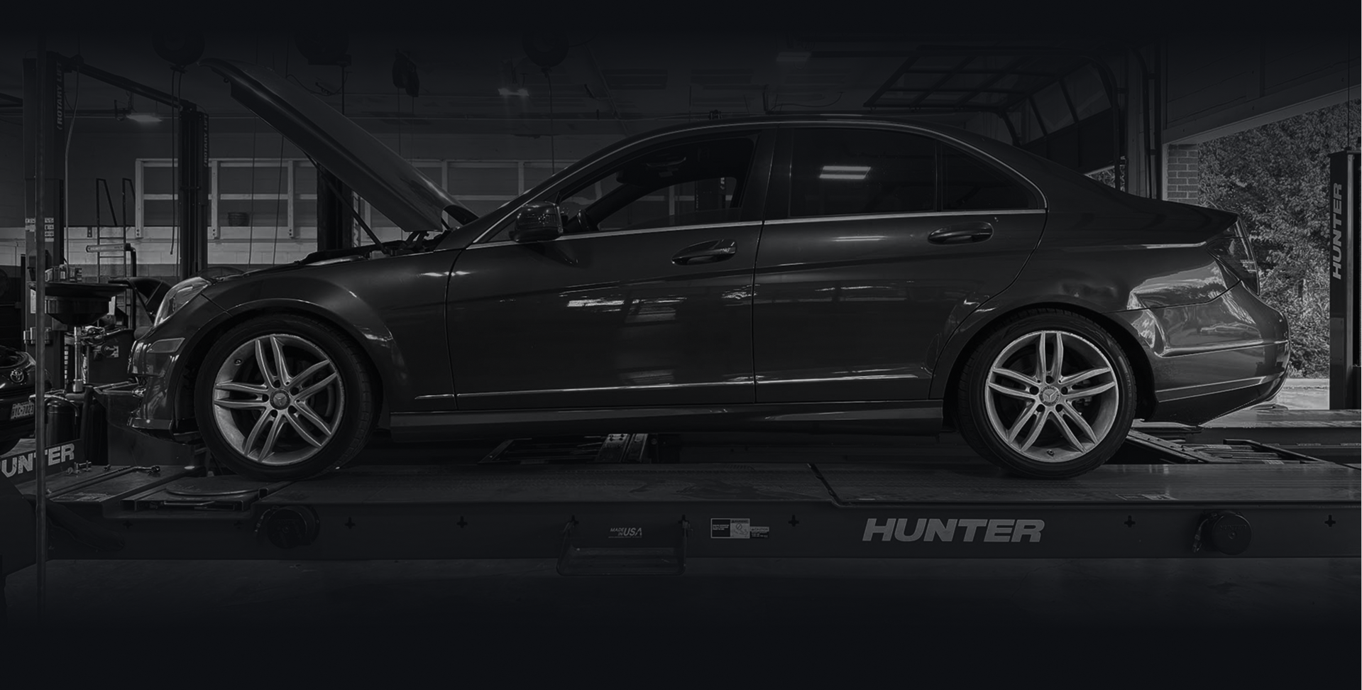 Black and white shot of a dark car on a Hunter alignment machine in a garage with the hood open. | Auto Check Sugar Land