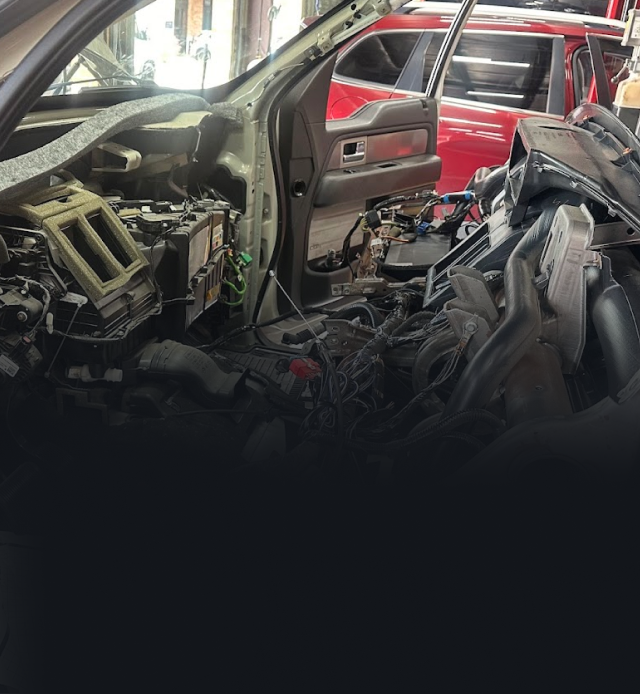 Car interior in a shop; dashboard and components removed, exposed wiring. Red car in the background.