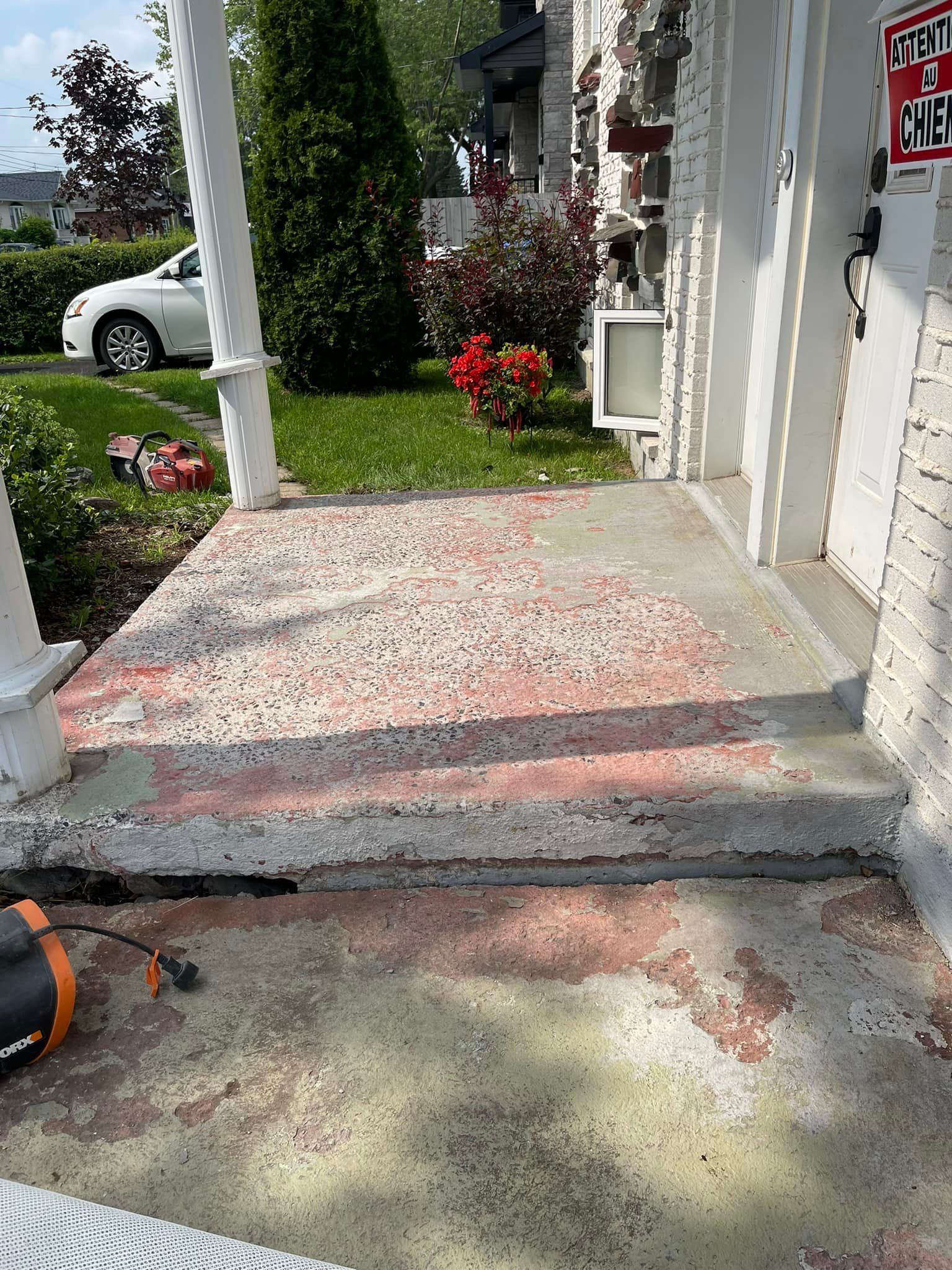 Porche en béton avec des traces de peinture rouge, menant à l'entrée de la maison. Comprend une marche et des piliers blancs.