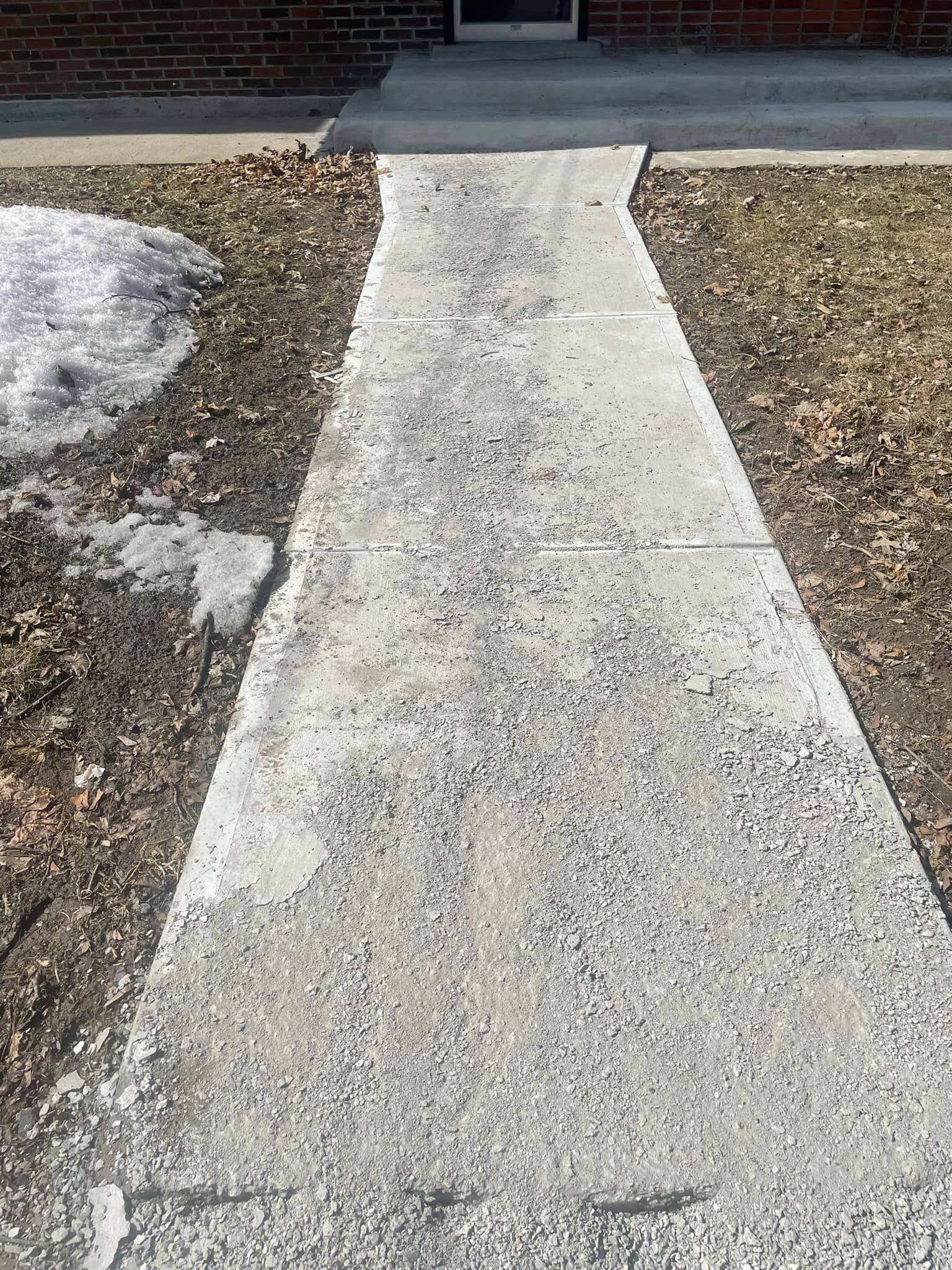 Un trottoir en béton mène de l'entrée de la maison, bordé de neige éparse et d'herbe.