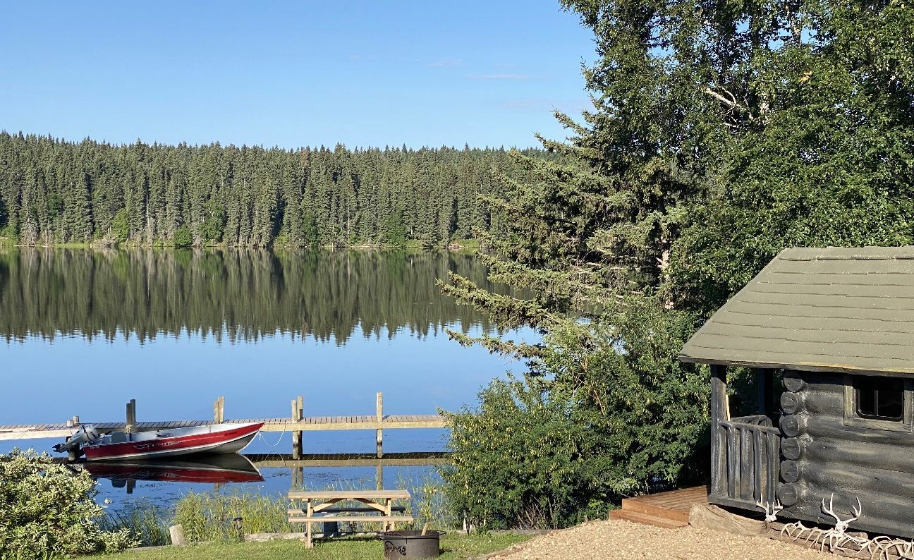 A log cabin is sitting next to a lake with a dock and a boat.