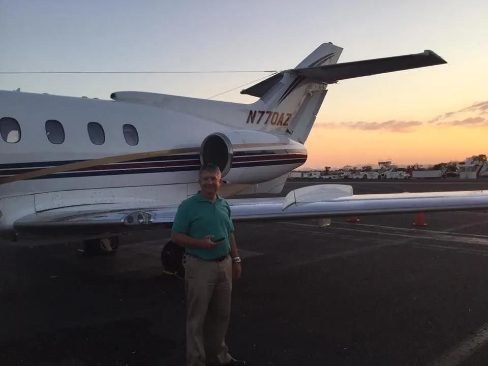 Man standing next to a private jet on a tarmac at sunset; plane is white with gold and blue stripes.