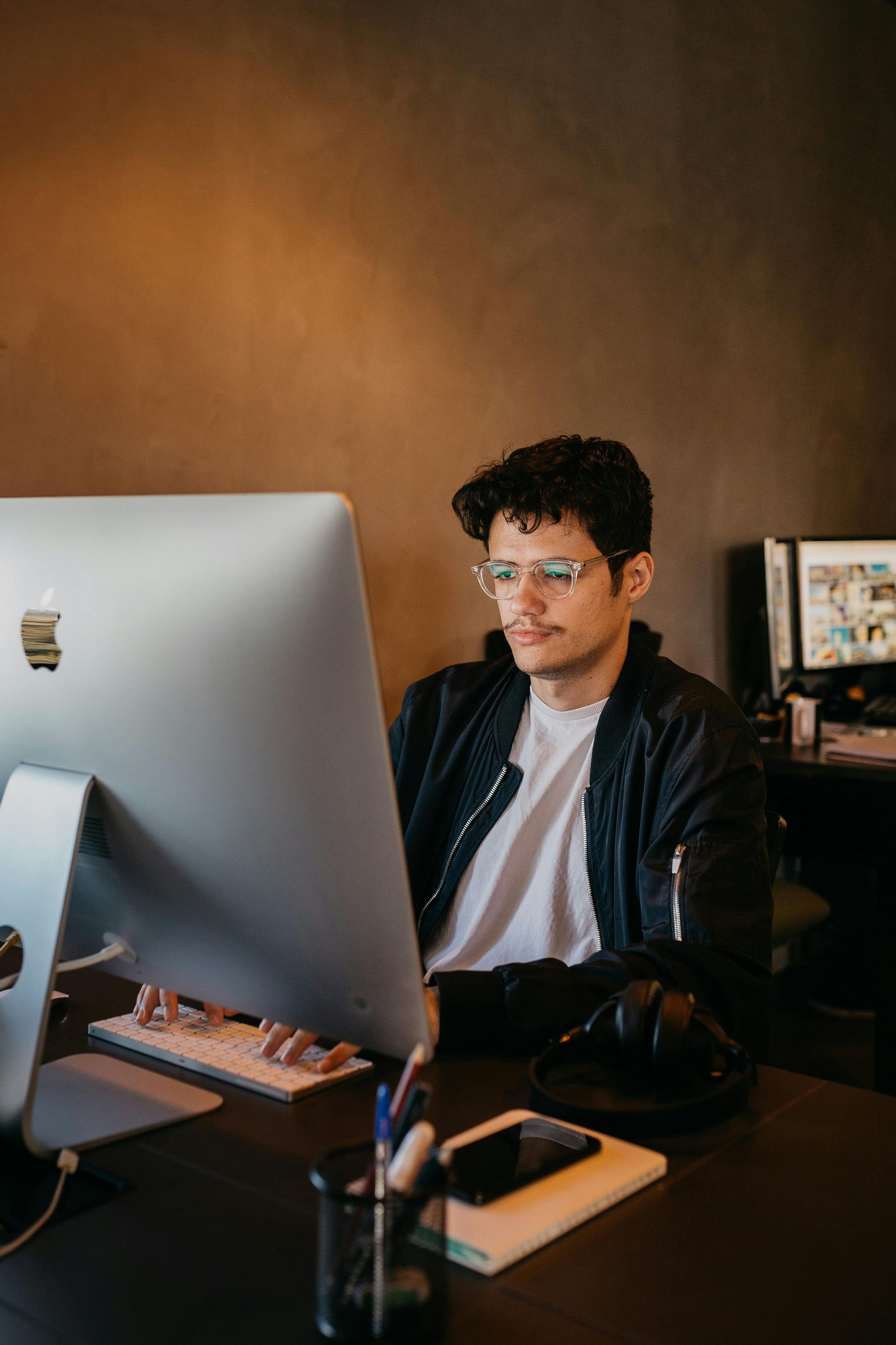 Man wearing glasses and a black jacket, seated at a desk with a desktop computer.