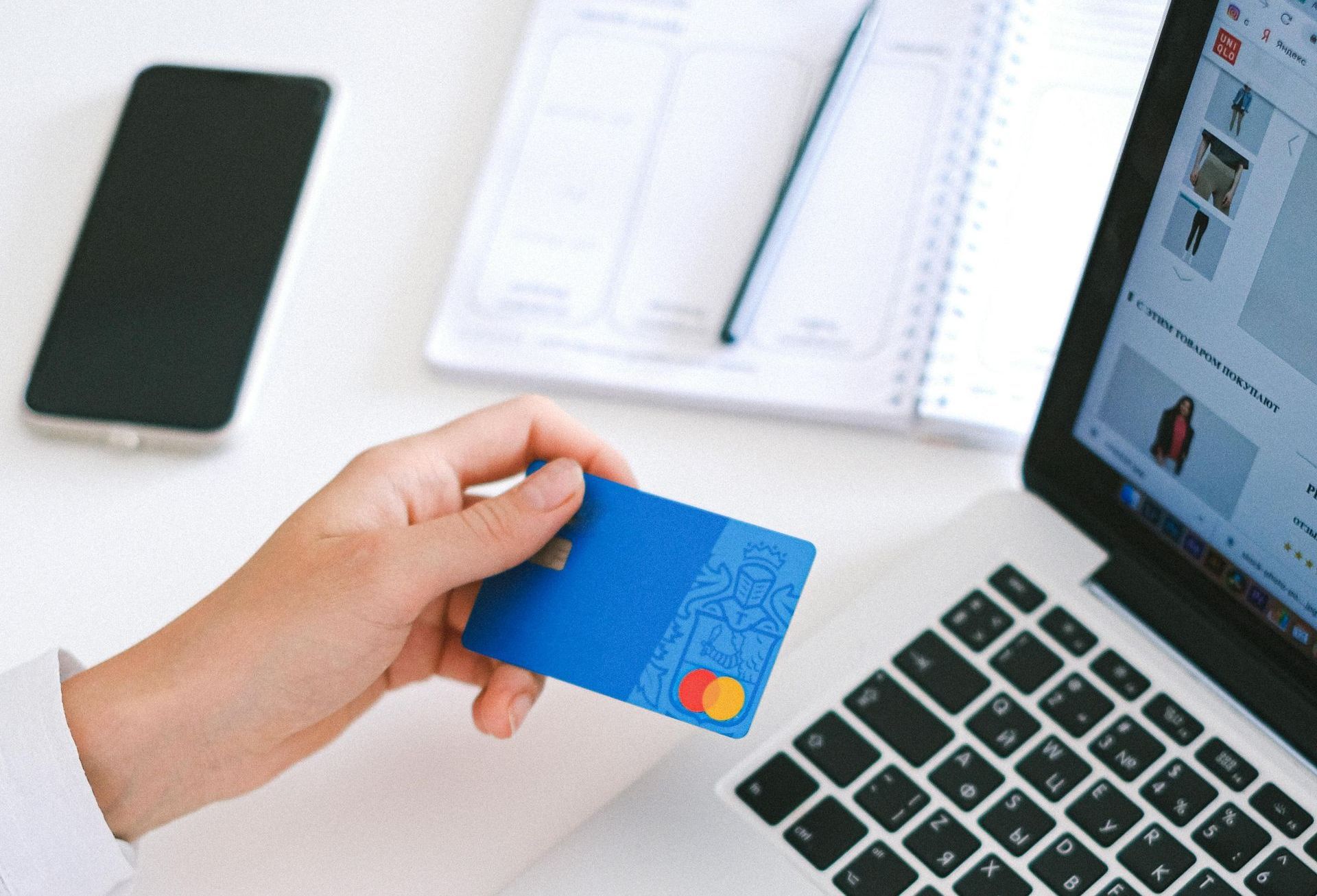Hand holding a blue credit card near a laptop displaying an online shopping website.