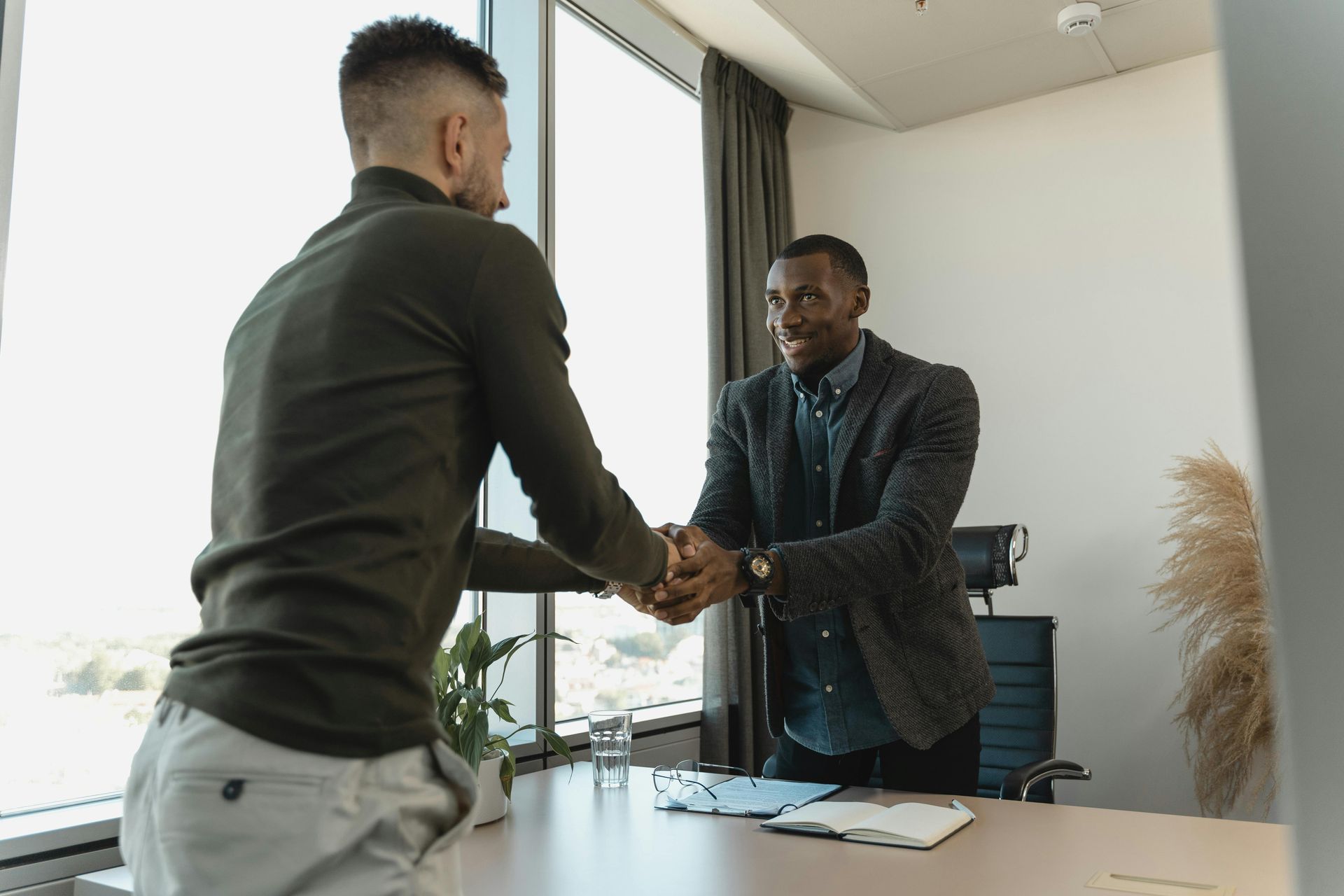 Two men shaking hands in a bright office; one in a blazer, the other in a sweater.