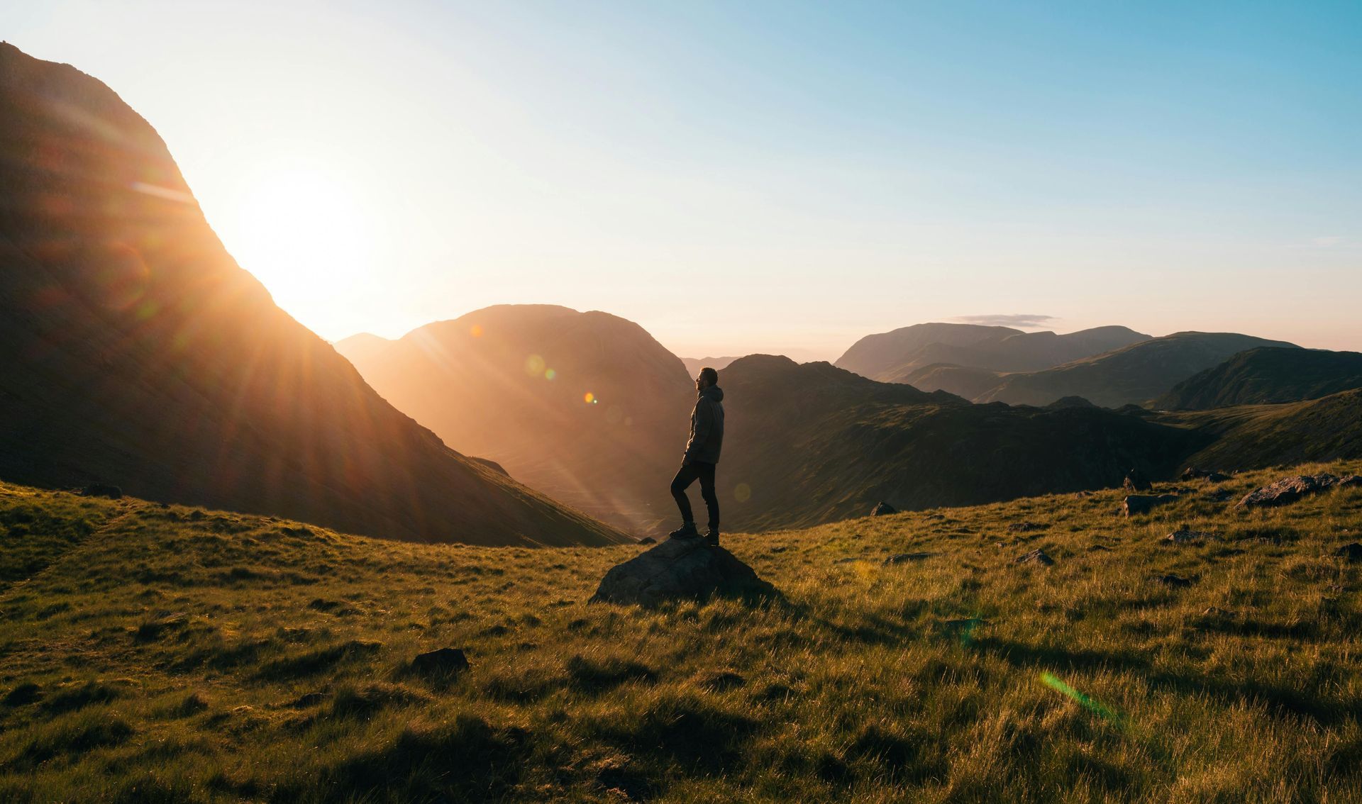 Person standing on a rock, silhouetted by the sun, overlooking mountains at sunset.