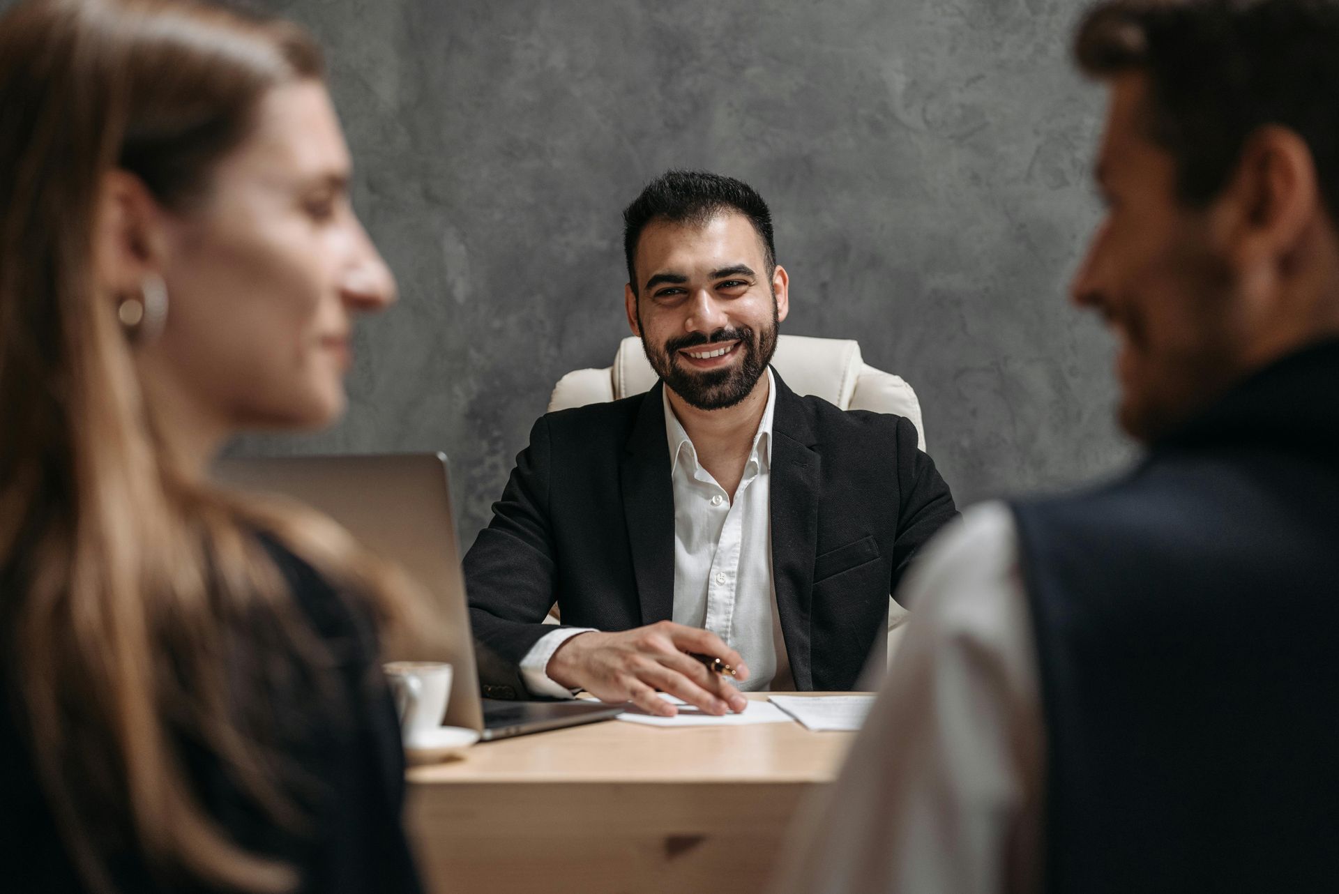 Person in a suit smiles at a couple. They sit at a desk with papers and a laptop in an office.