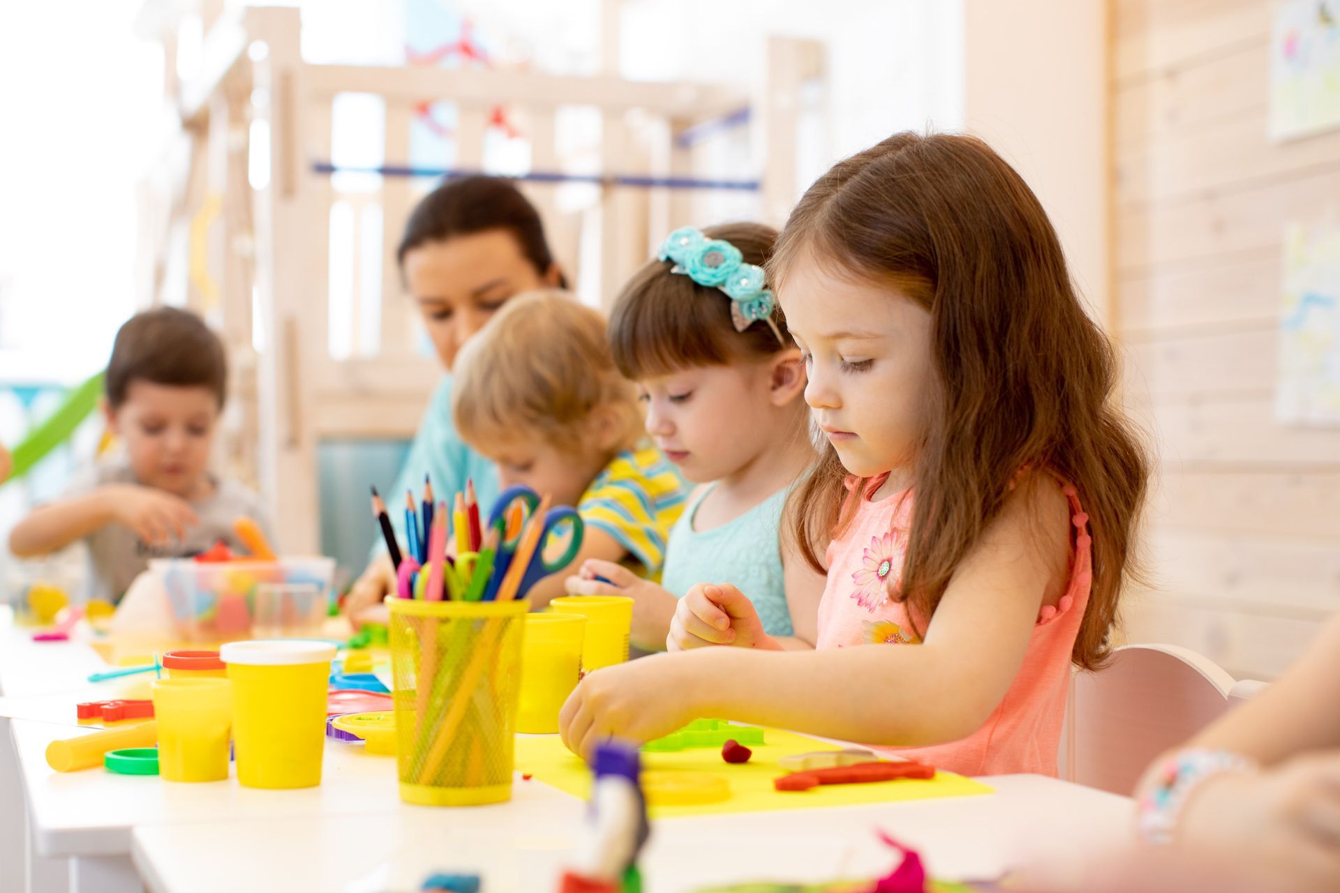 Children sit at a table in a bright classroom working on craft projects with art supplies and clay.