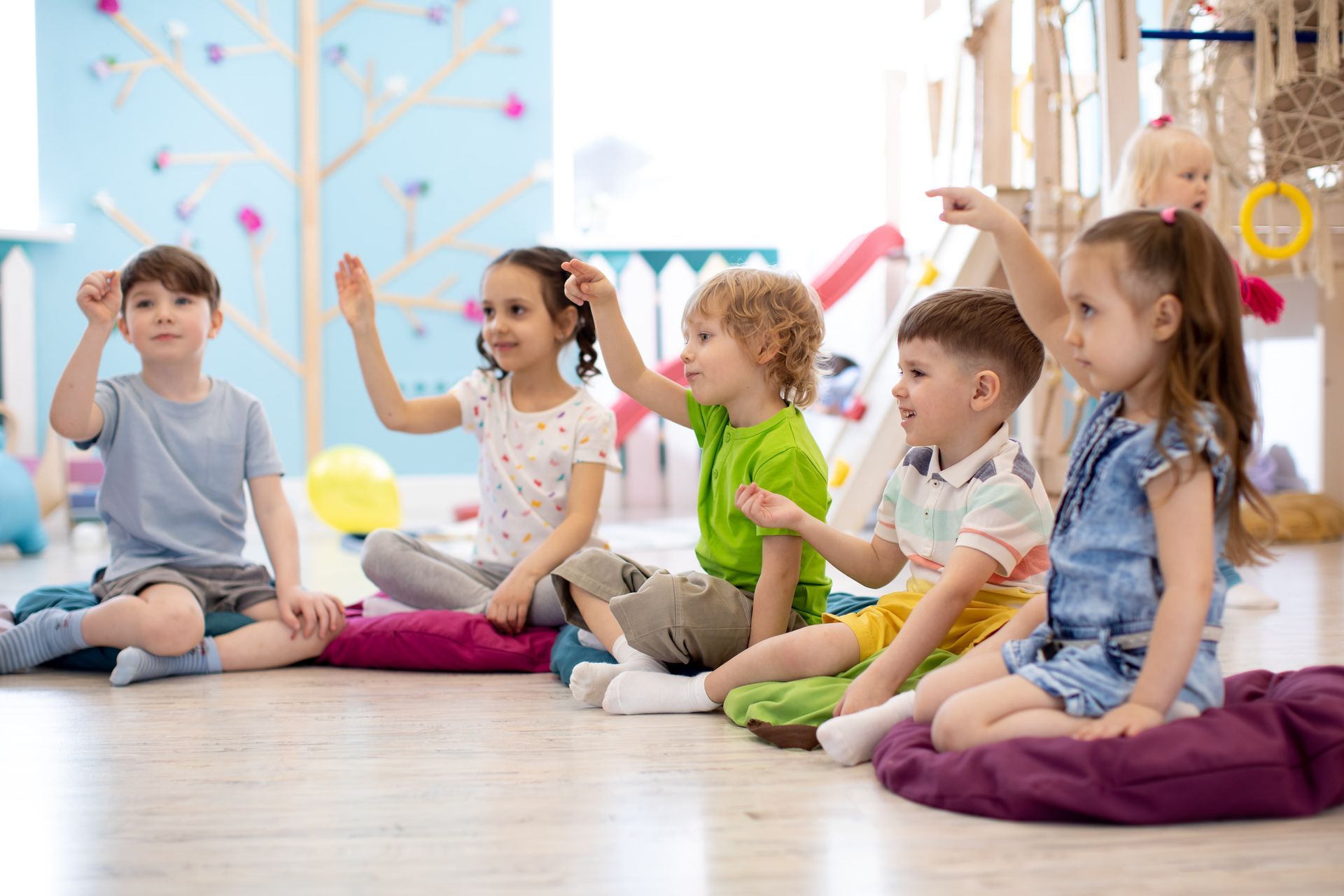 A group of children sitting on the floor in a colorful classroom, with some raising their hands to participate.