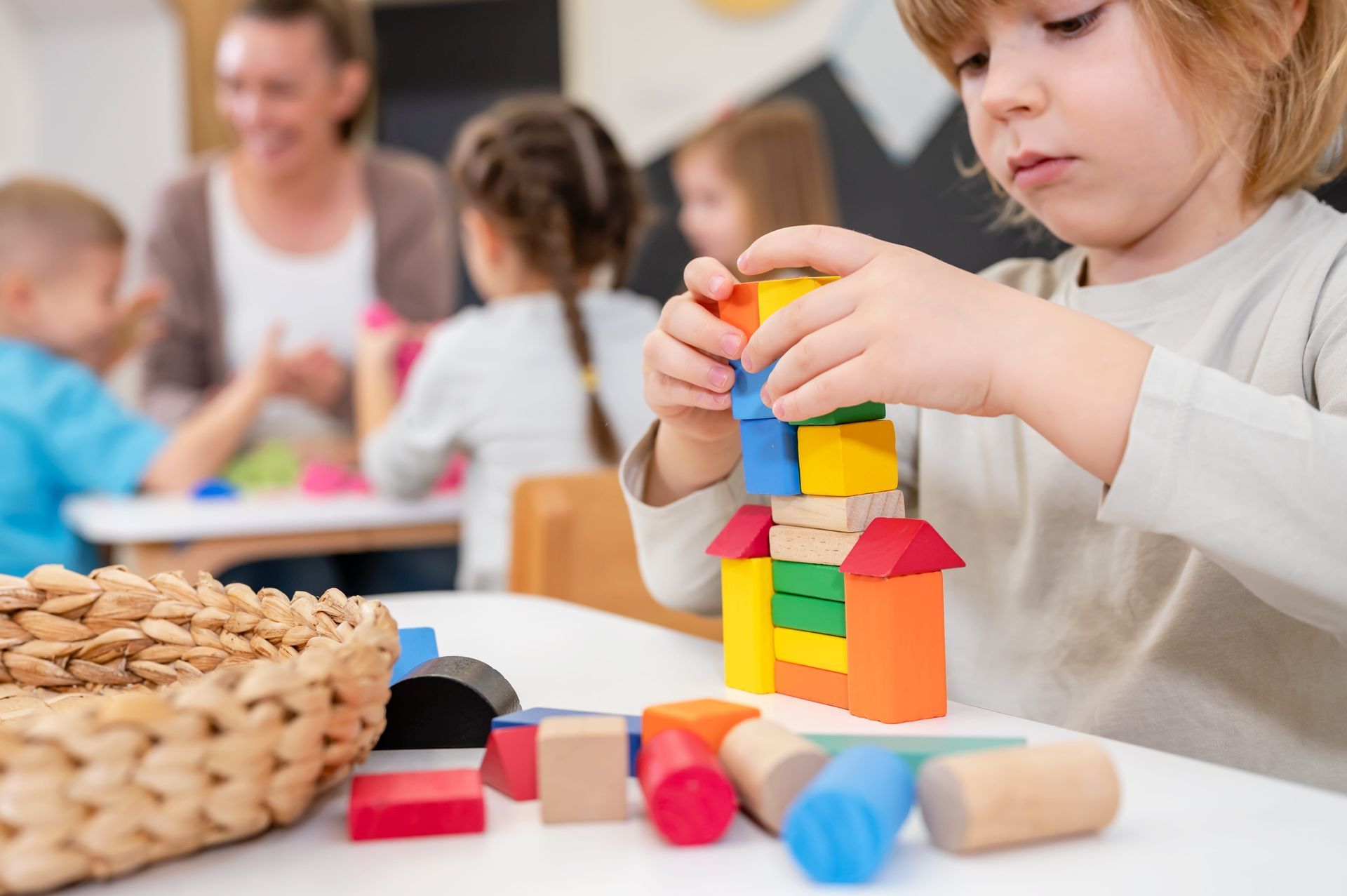 A young child in a classroom builds a tower with colorful wooden blocks, with other children and a teacher in the blur.
