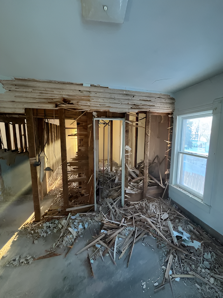 Room under renovation with exposed framing, debris on the floor, and damaged drywall near a window.