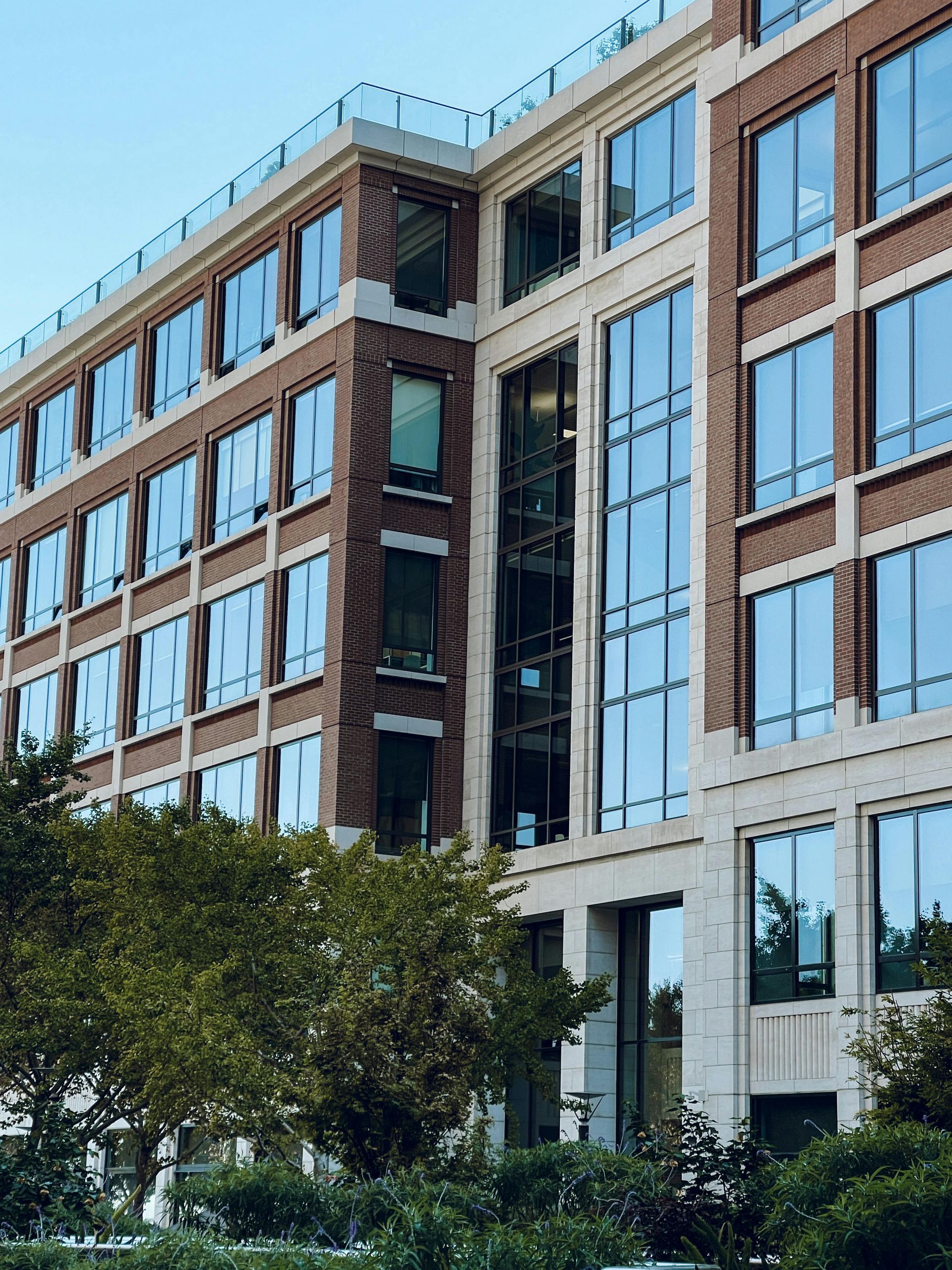Modern multi-story office building with brown brick and white panels beside leafy trees under a clear blue sky