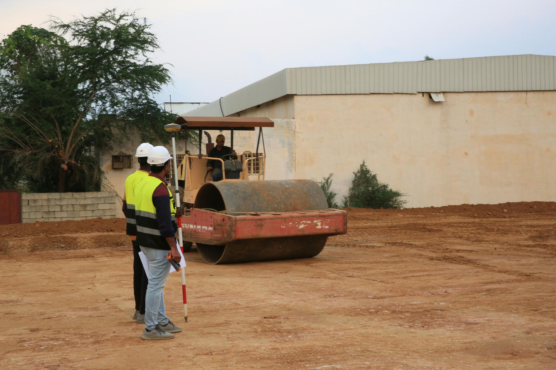 Worker in safety vest guiding a road roller on a dirt lot beside a beige building.