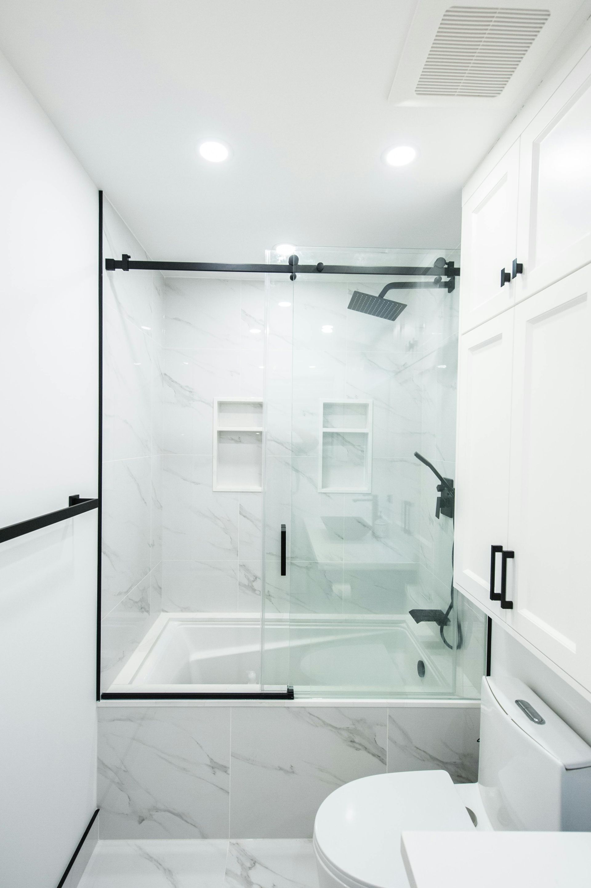 Modern white bathroom with glass shower, black fixtures, toilet, and bright ceiling lights