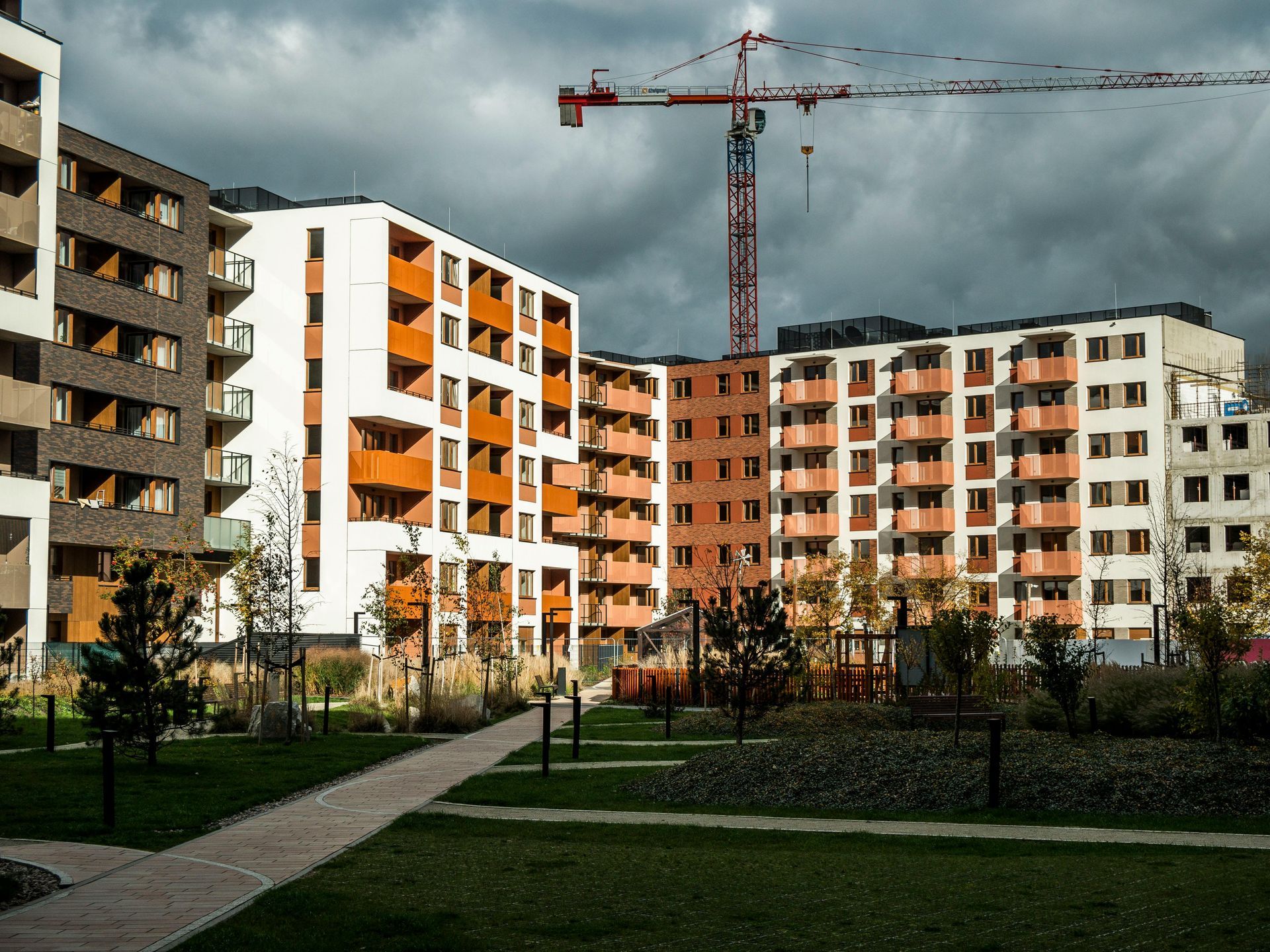 Apartment buildings under construction beside a grassy path, with a crane and cloudy sky.