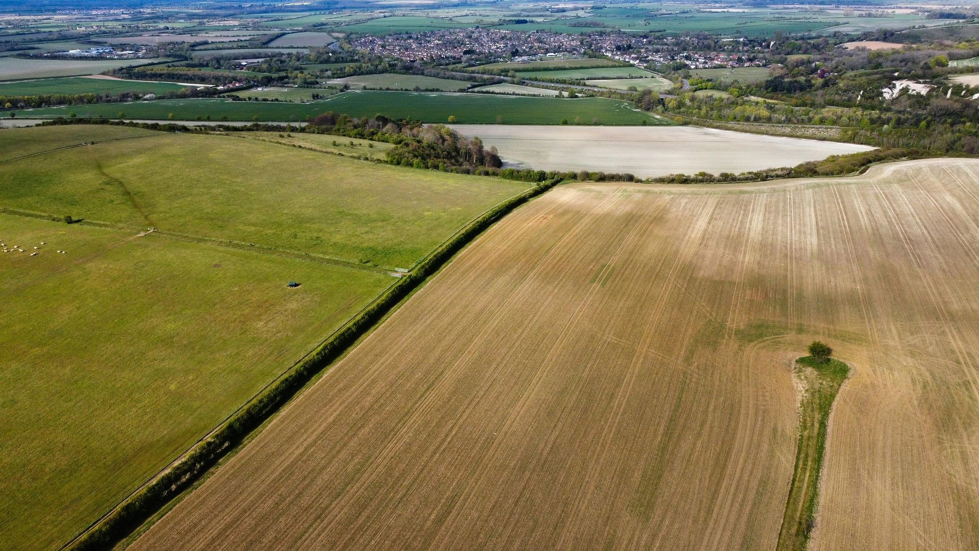Aerial view of green and tan farmland with a pale water reservoir and distant village under a cloudy sky