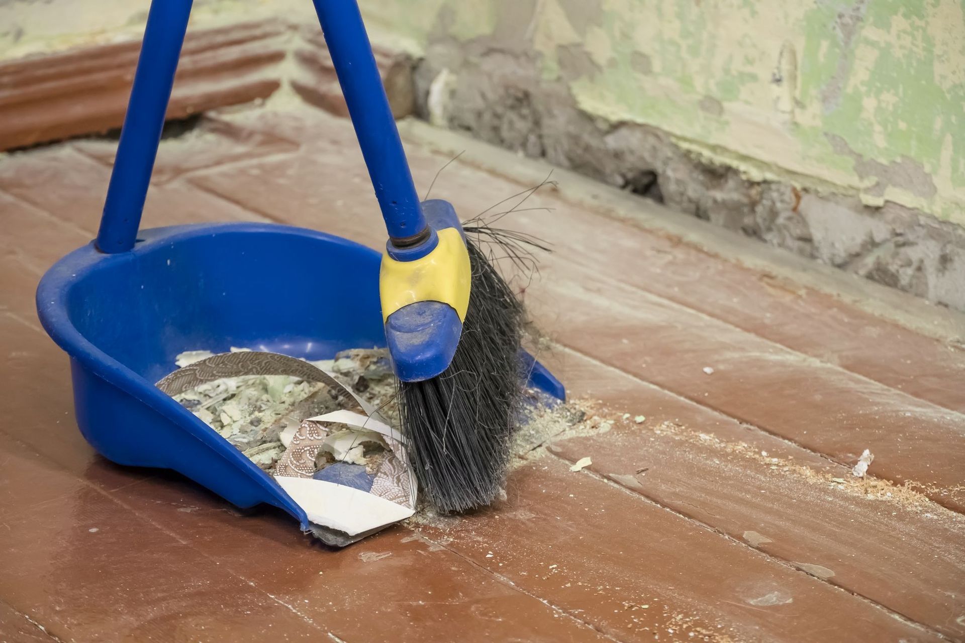 Blue dustpan and broom sweeping debris on a dusty floor near a wall.