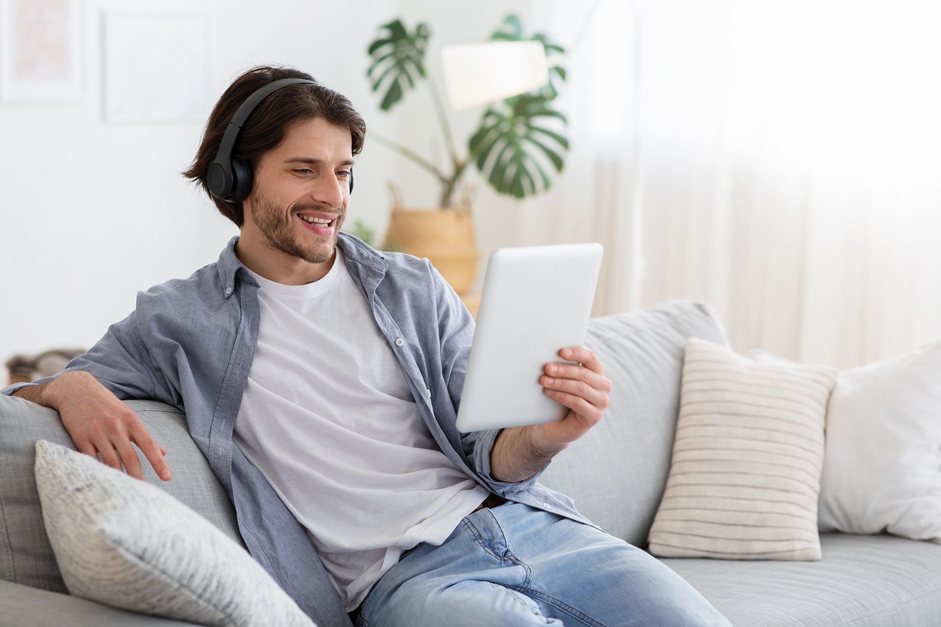 Hombre con auriculares, sonriendo, mirando una tableta mientras se relaja en un sofá en una sala de estar luminosa.