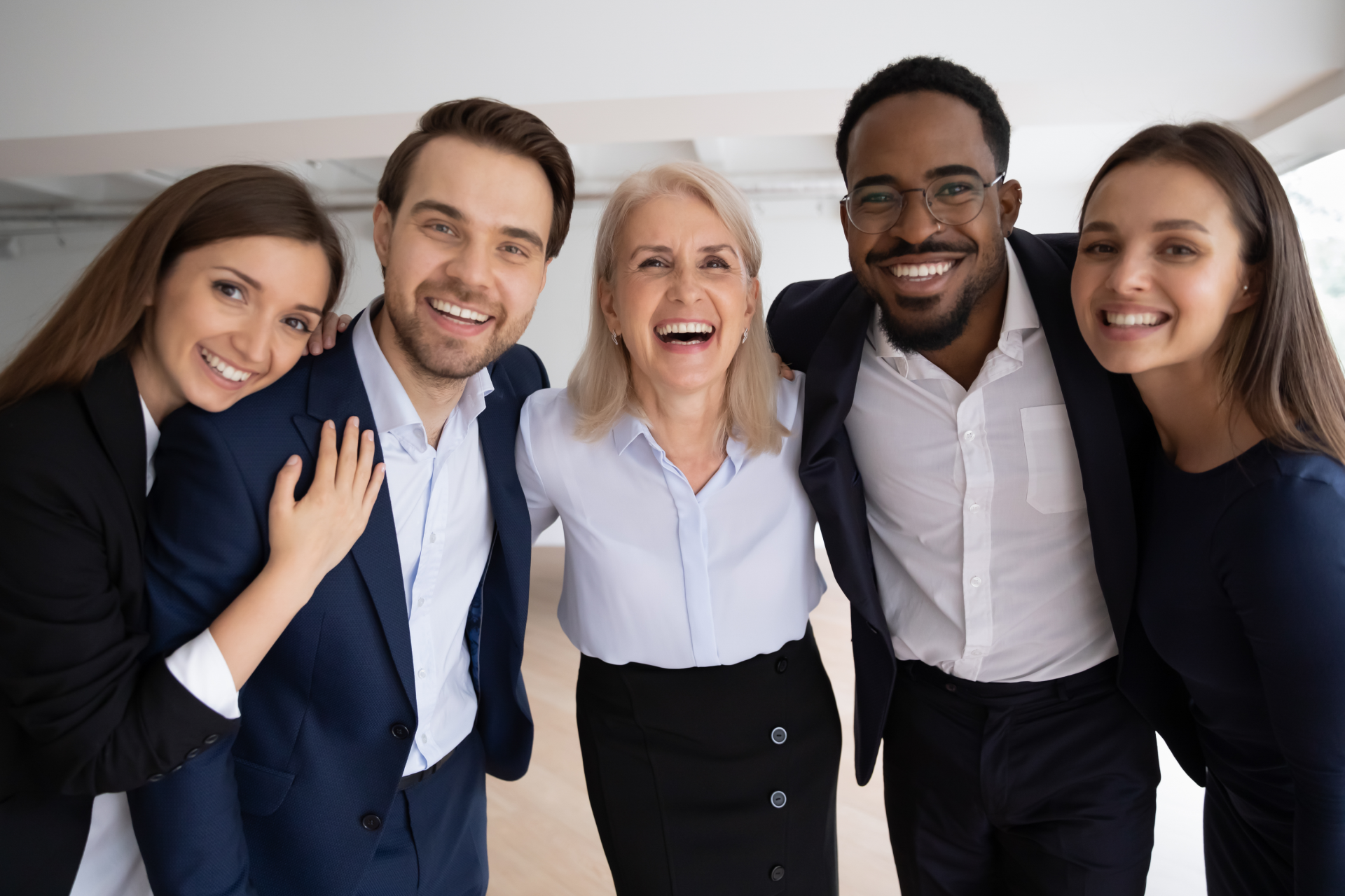 Five colleagues stand in a close group, smiling broadly and looking at the camera in a bright, modern office space.