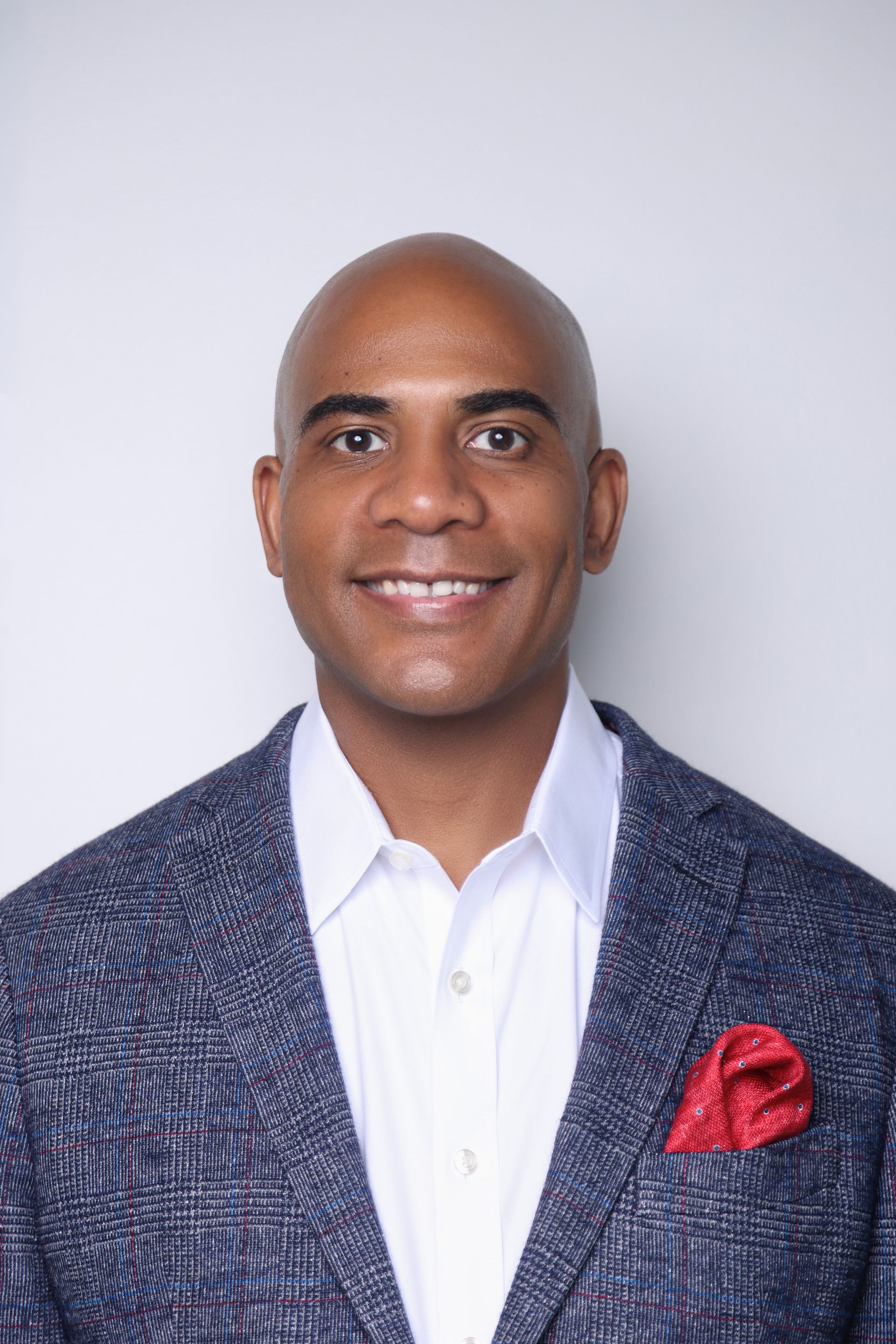 A smiling man with a shaved head wears a white collared shirt, a textured blue blazer, and a red pocket square.