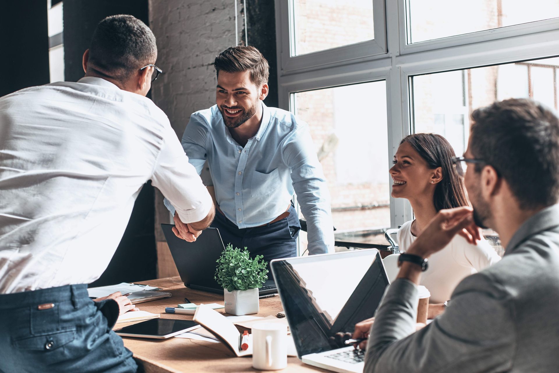 Business professionals shaking hands across a table during a meeting in a bright, modern office.