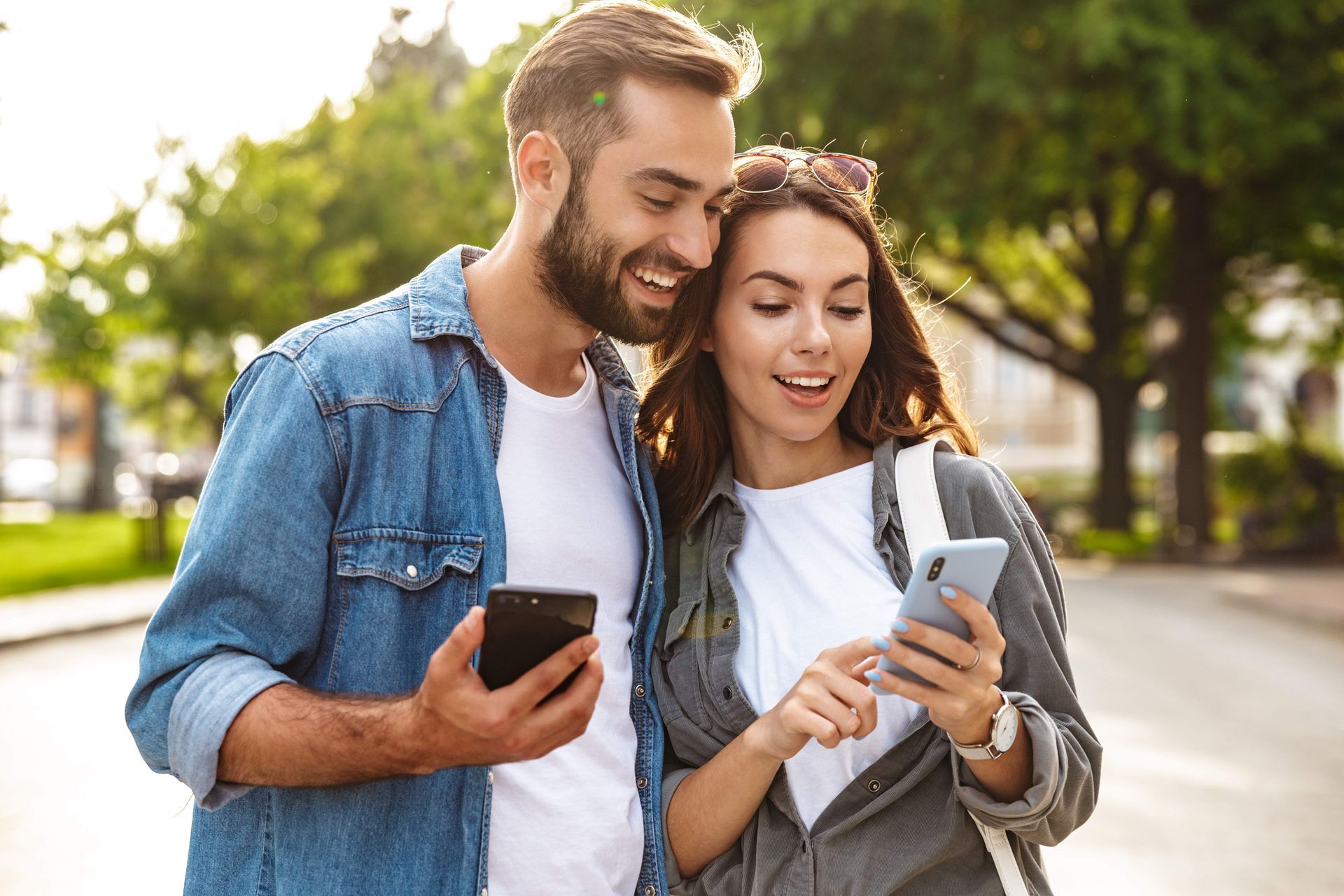 A couple smiling while looking at their smartphones together in an outdoor park setting.